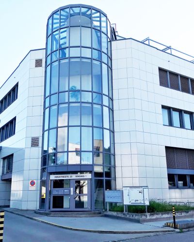 Unser neuer Standort in Oberentfelden. Das Bild zeigt die Außenansicht eines großen dreistöckigen Industriegebäudes. Das markante Treppenhaus mit Glasfenstern ist deutlich erkennbar.